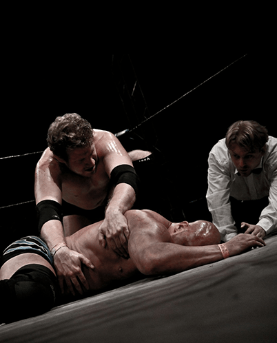 A wrestler holds an opponent down on the mat, while a referee observes closely. The scene captures the intensity of a professional wrestling match.